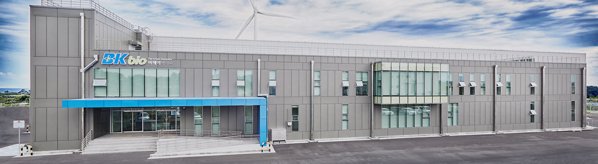 BK bio's Jeju factory exterior in the Lava-Seawater Industrial Complex, signage and a wind turbine against the Jeju sky.